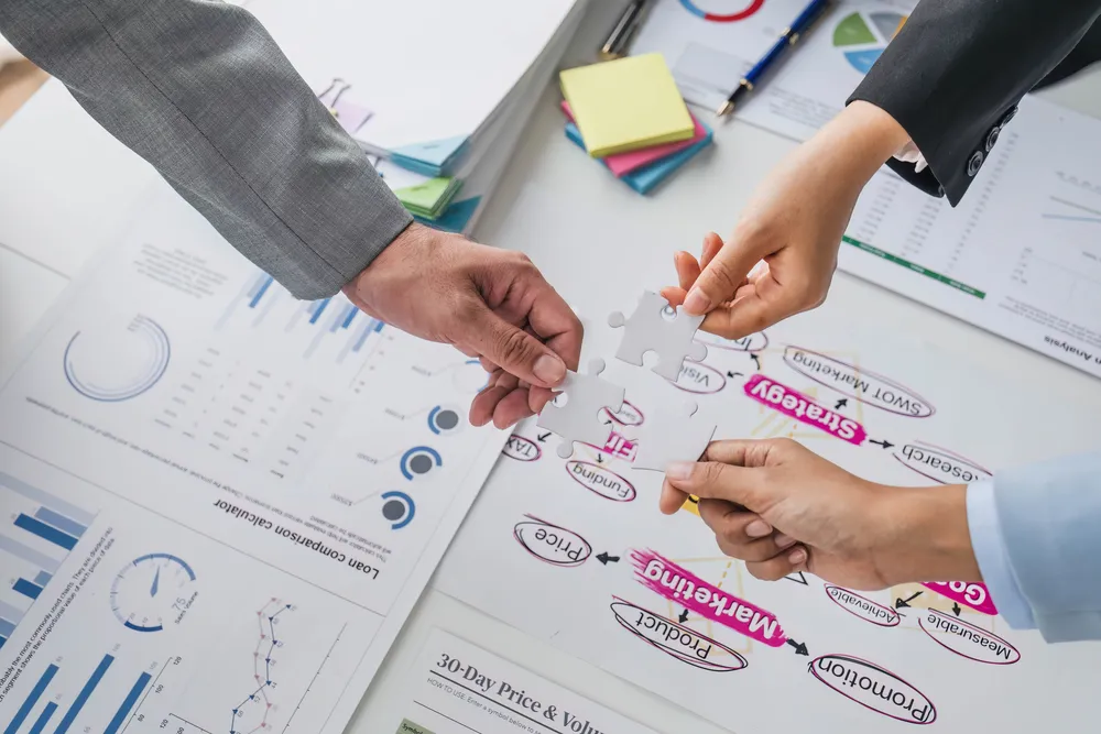 Tres manos uniendo piezas de rompecabezas sobre diagramas de estrategia kaizen y reportes de desempeño.